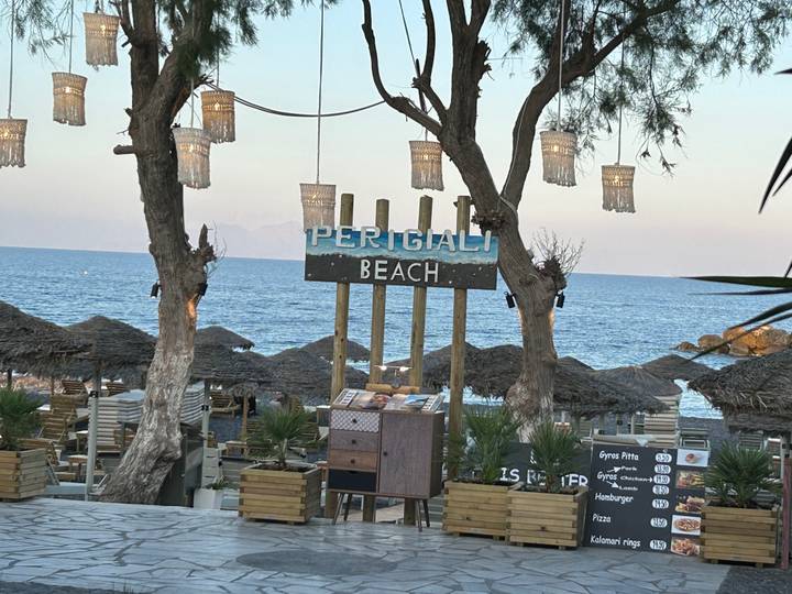 Beachfront restaurant sign reading 'Perigiali Beach' with thatched umbrellas and blue sea beyond.