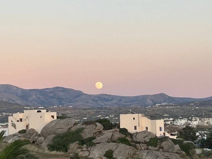 Rising full moon over pastel sky above Cycladic white houses and rocky hills.