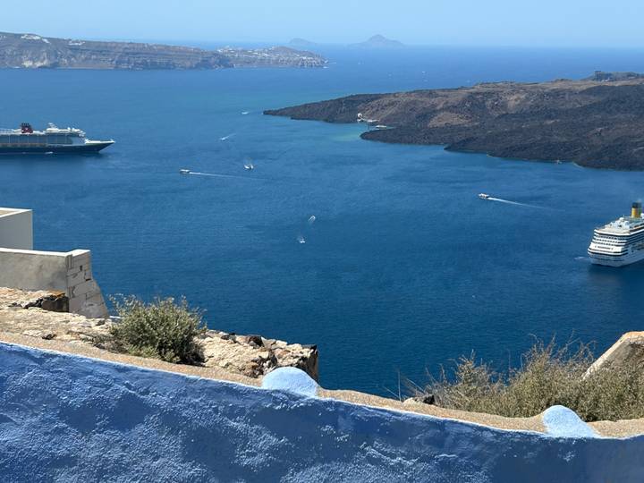 Deep blue caldera waters with cruise ships and speedboats viewed from whitewashed Santorini cliff.