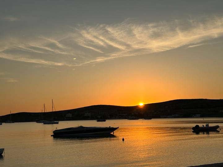 Sunset over a calm bay with small boats silhouetted against an orange sky and low rolling hills.