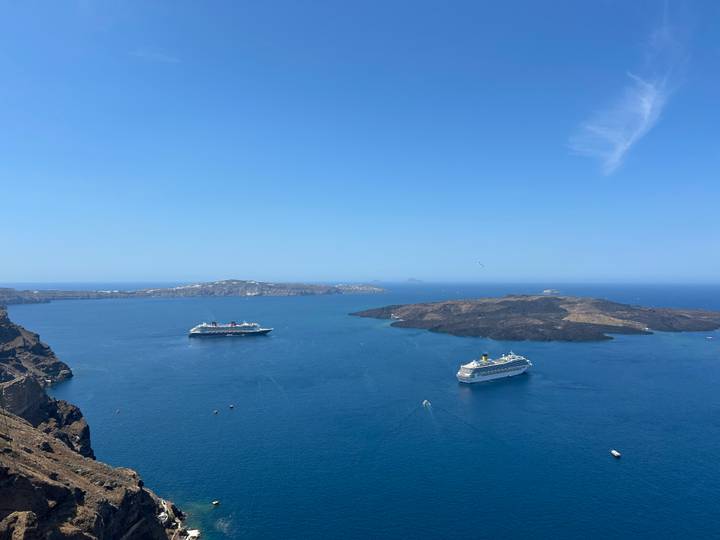 Wide aerial view over Santorini caldera with cruise ships anchored in deep blue waters beneath a clear sky.