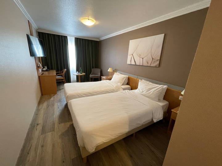Simple hotel room with twin beds, wood furniture and a tall window with dark curtains.