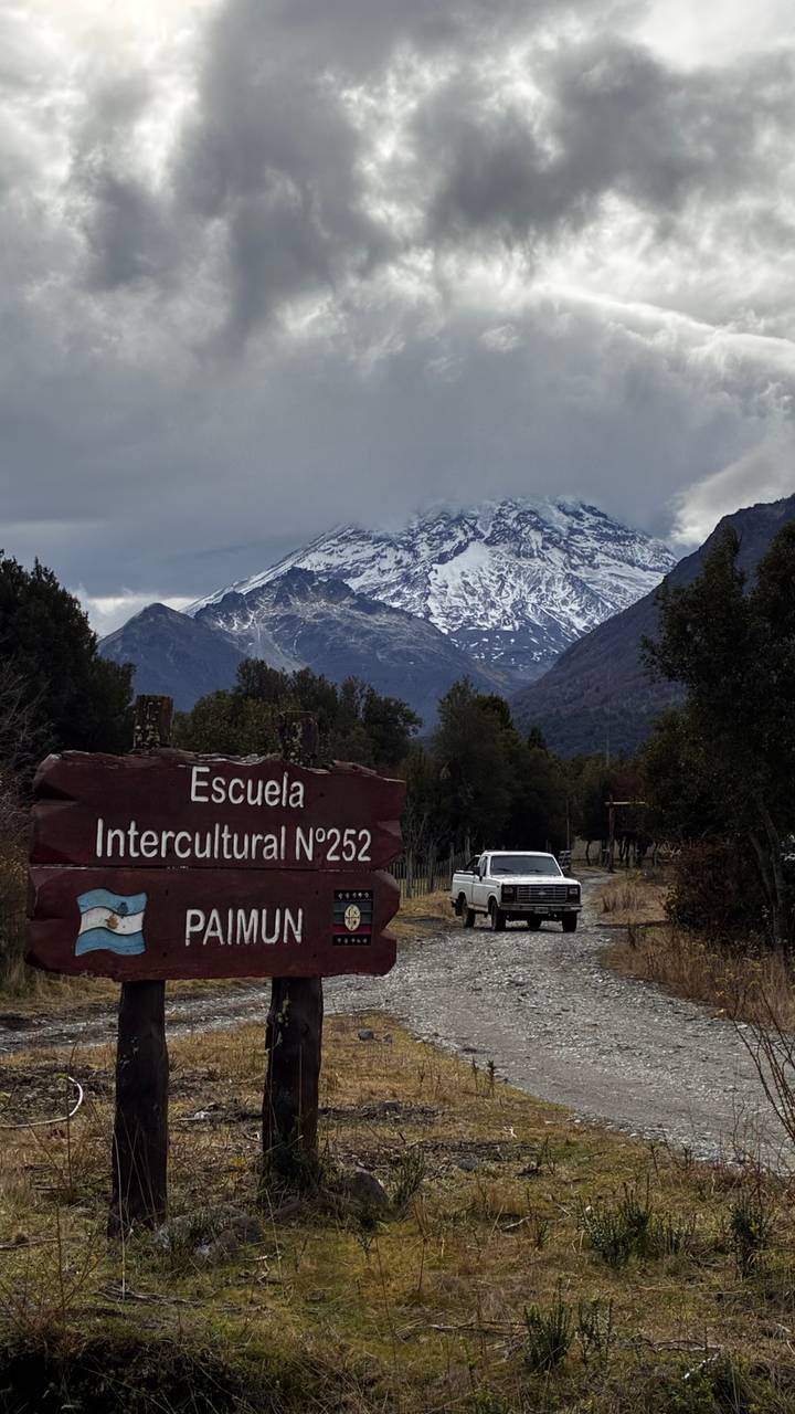 Rustic wooden sign for Escuela Intercultural N°252 with cloud-covered snowcapped mountain behind.