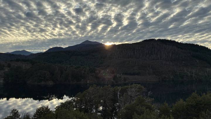 Morning sun peeking over a mountain ridge reflecting on a calm forest lake under patterned clouds.