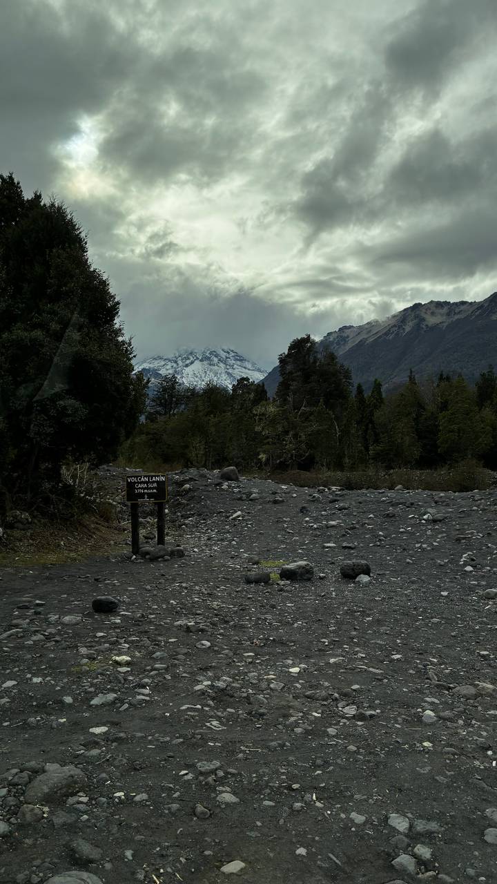 Rocky riverbed with sign indicating Volcán Lanín trail and snow-capped peak partially obscured by clouds.