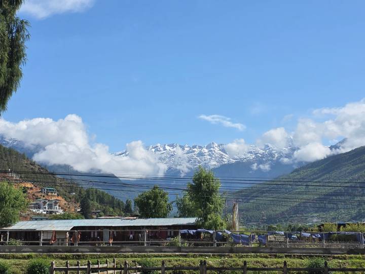 Snow-capped Himalayan peaks rise above a green valley dotted with houses and framed by a clear blue sky.