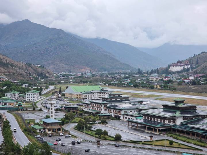A small mountain airport runway with surrounding town and monastery set in a green valley under low clouds.