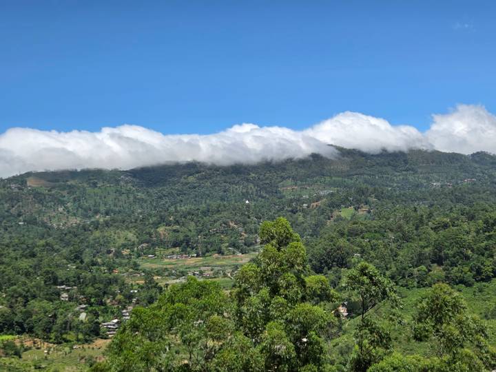 Verdant mountains rise above a valley town as low clouds drift along the ridgeline under a clear blue sky.