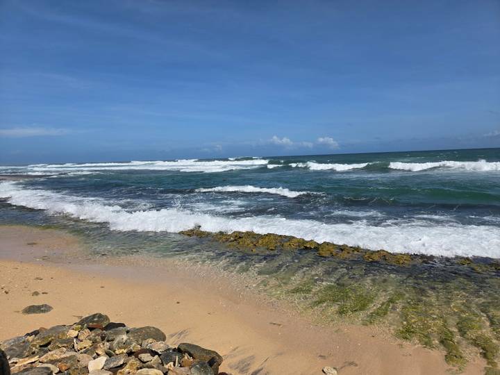 Waves crash onto a sandy tropical beach with clear aqua shallows under a bright blue sky.