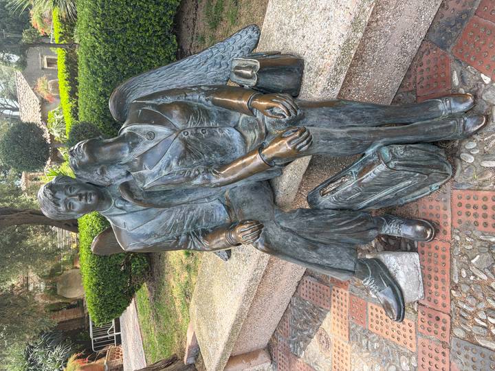 Bronze sculpture of a suited man and woman with wings resting together on a stone bench in a shaded garden