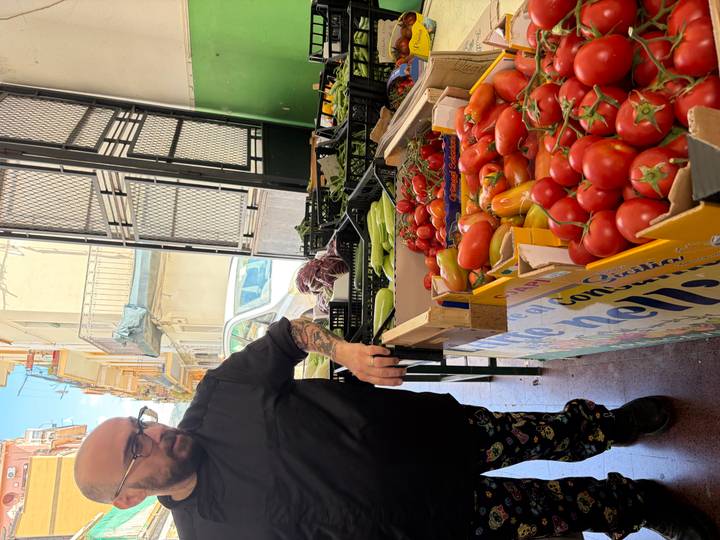 Tattooed vendor standing beside crates of bright red tomatoes and fresh produce inside an open market