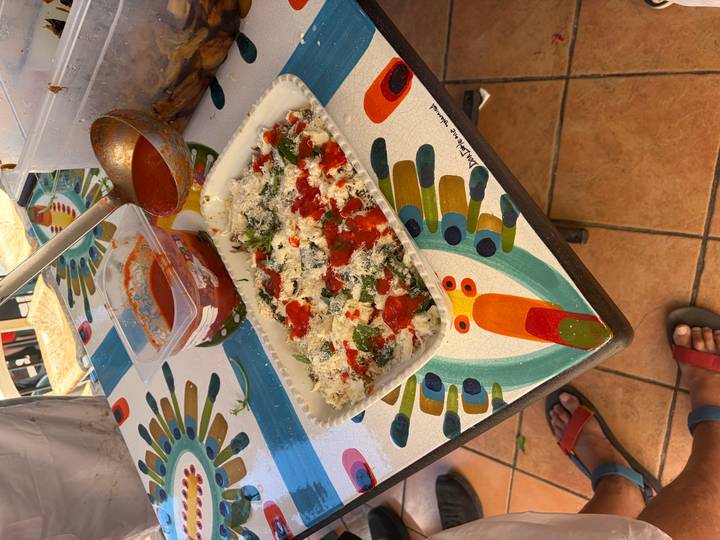 Tray of lasagna being assembled with tomato sauce and ricotta on a colourful tiled table