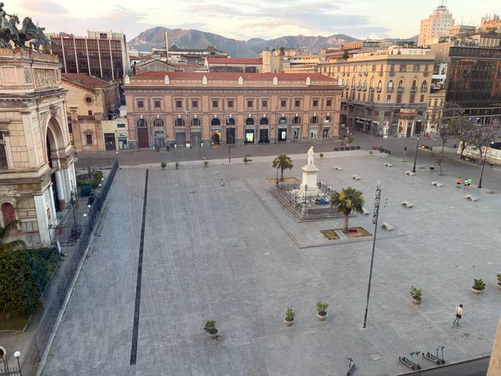 Wide shot of a spacious stone plaza with palm trees, benches and neoclassical buildings at dusk