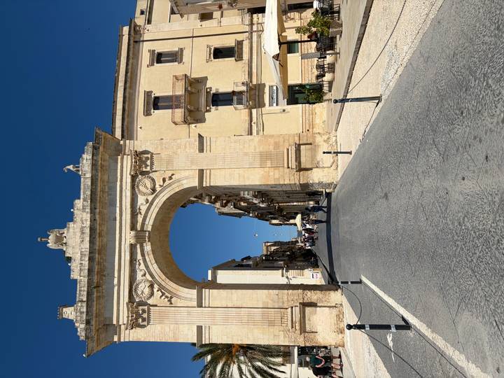 Grand stone triumphal arch leading into a historic street lined with baroque buildings under clear skies