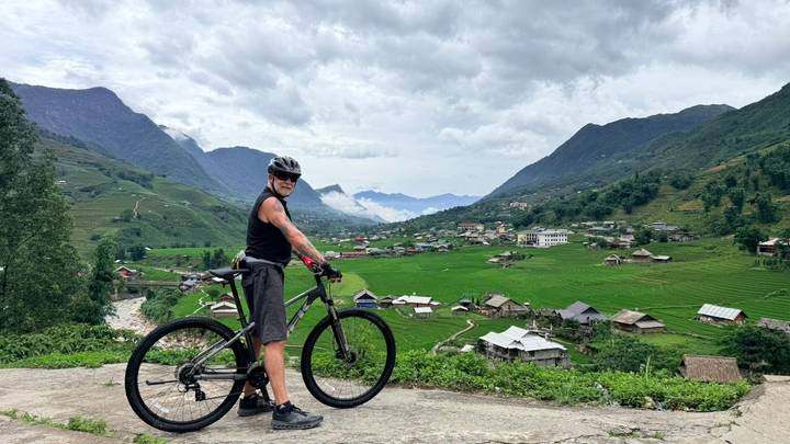 Cyclist poses with mountain bike overlooking a lush valley of rice fields and distant peaks under cloudy skies