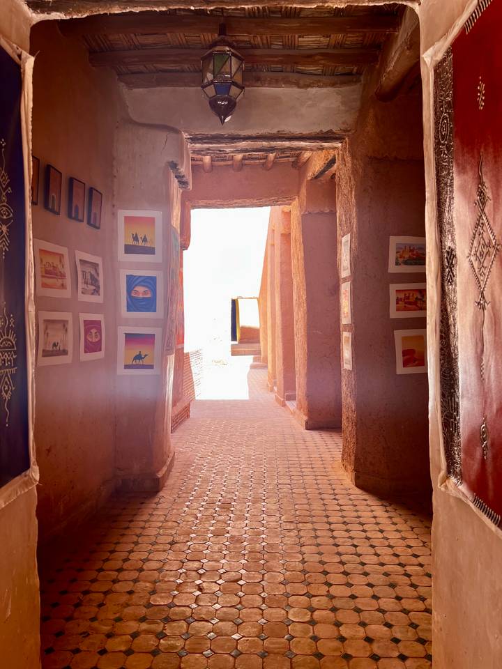 Sunlit narrow adobe corridor decorated with framed desert photography prints leading to a bright doorway