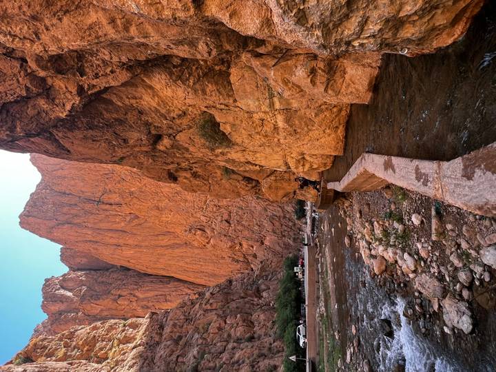 Steep reddish canyon walls tower above a shallow river and walking path in a dramatic gorge