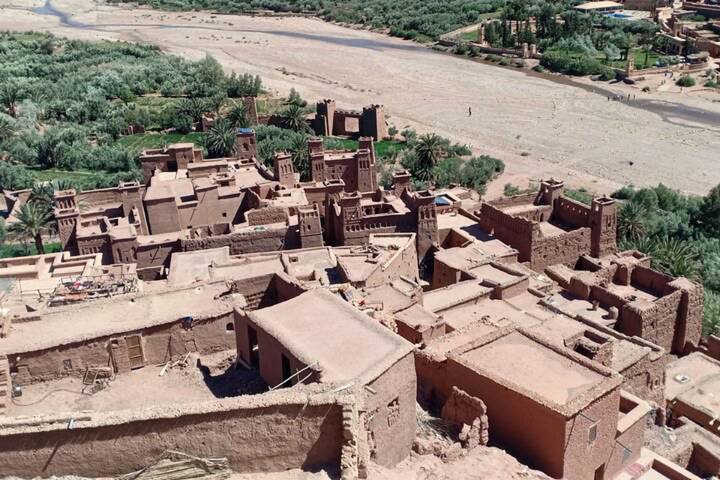 Aerial view of the earthen fortifications and date palms of Ait Benhaddou beside a dry riverbed.