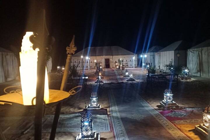Nighttime desert camp illuminated by lanterns and rugs leading to canvas tents.