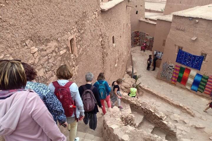 Tourists descend narrow stone steps between adobe walls lined with colorful textiles.