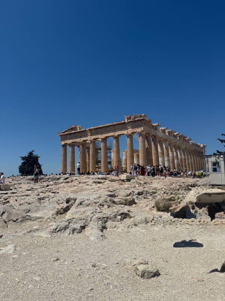 The Parthenon stands majestic on the Acropolis as crowds explore the ancient marble ruins against a deep blue sky.