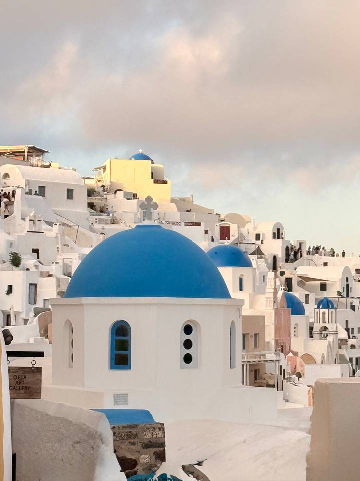 Iconic whitewashed buildings topped with vivid blue domes crowd the cliffs of Oia in soft evening light.