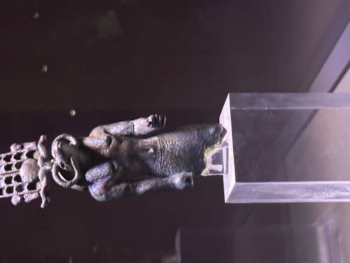 Small bronze Egyptian deity statue displayed on a clear pedestal inside a museum exhibit