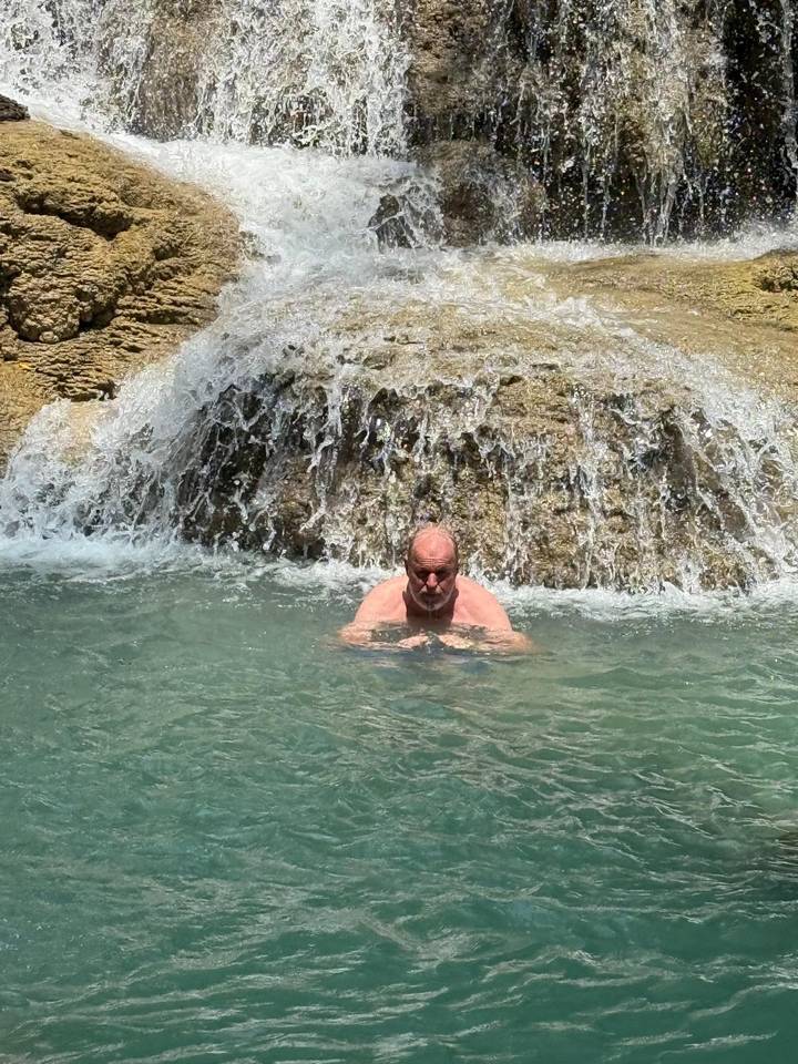 Older man cooling under a small waterfall in turquoise natural pool.