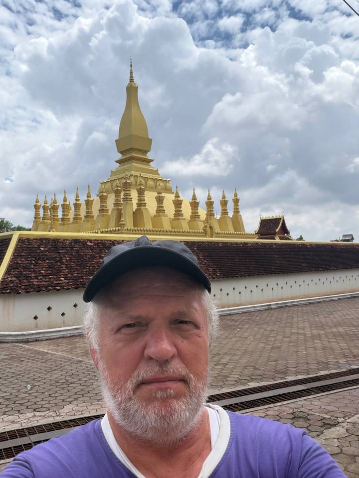 Selfie with golden stupa of Pha That Luang rising behind tiled wall in cloudy sky.