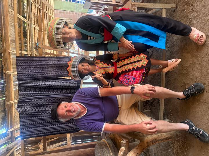 Tourist seated with two local people in traditional hill-tribe clothing inside wooden shelter.