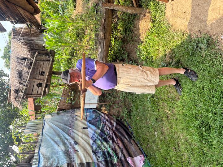 Older man aiming a wooden crossbow in a rural garden with maize plants.