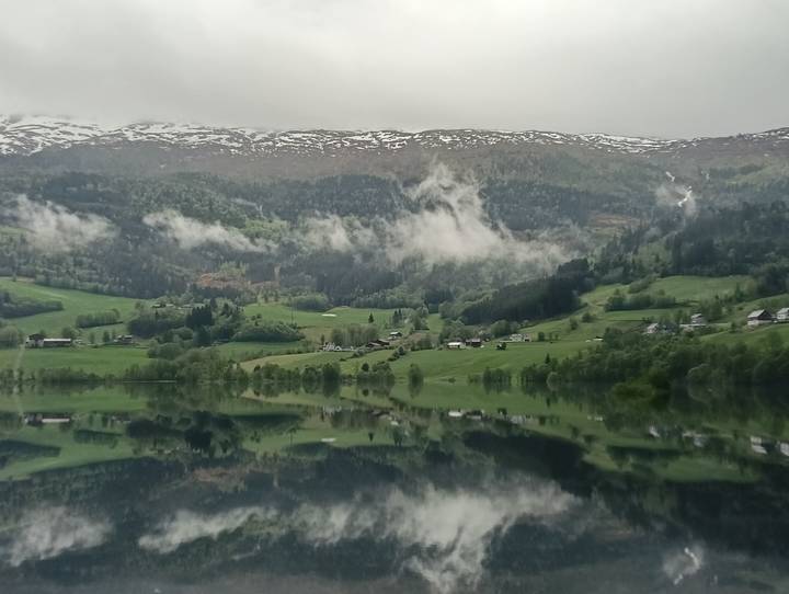 Mirror-calm fjord reflects lush green slopes and low clouds beneath snow-dusted ridge.