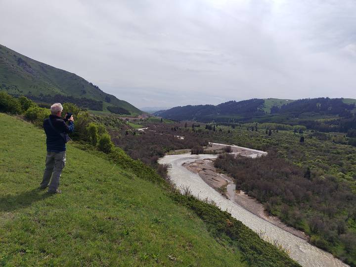 Man taking a photo of a winding river valley surrounded by green hills and distant snow-capped mountains.