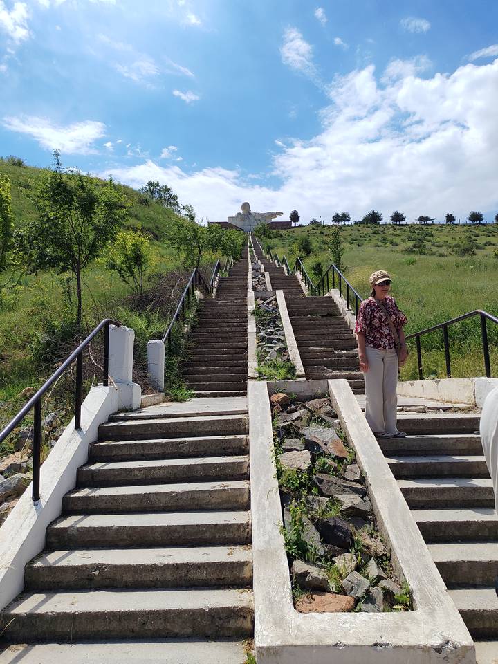 Long concrete stairway climbing a grassy hillside toward a large white statue with outstretched arms, with a woman posing near the bottom.