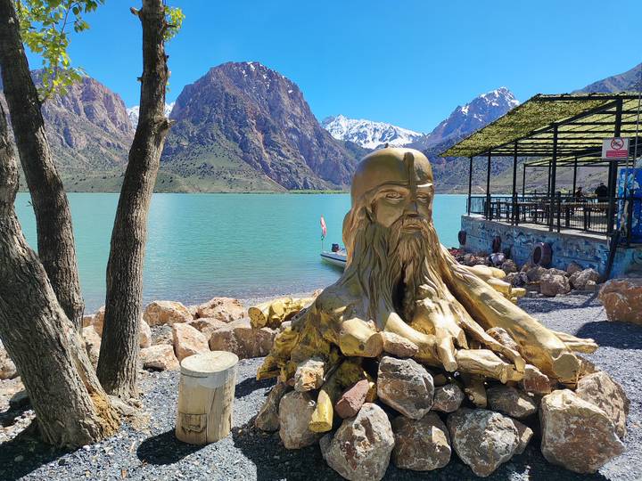 Golden sculpture of a bearded head beside a turquoise lake framed by dramatic snow-tipped mountains.