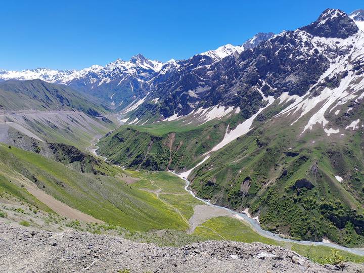Wide alpine valley with lush green slopes, snow-capped peaks and patches of late-season snow under a clear blue sky.