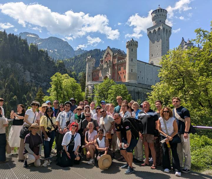 Tour group gathered in front of the fairytale Neuschwanstein Castle surrounded by alpine forest.