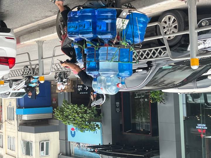 Motorcyclist transporting stacks of large blue water bottles along a busy city street.