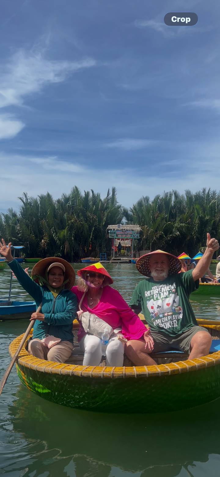 Three smiling tourists in conical hats give thumbs-up while riding a round basket boat on calm water fringed with palm trees.