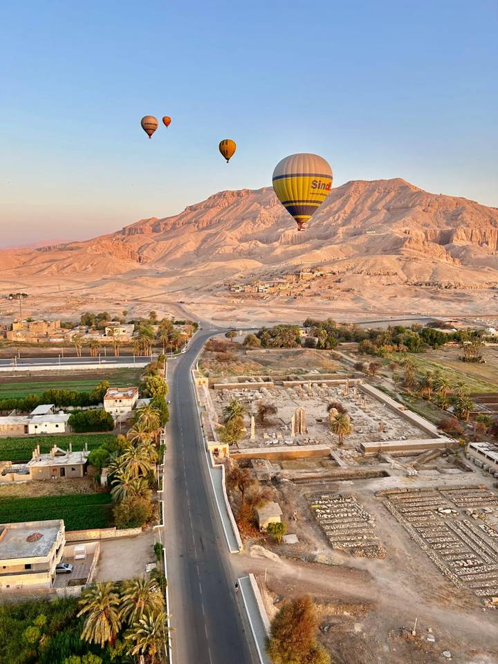 Hot-air balloon floating above desert landscape dotted with ancient ruins and farms in warm sunrise light.