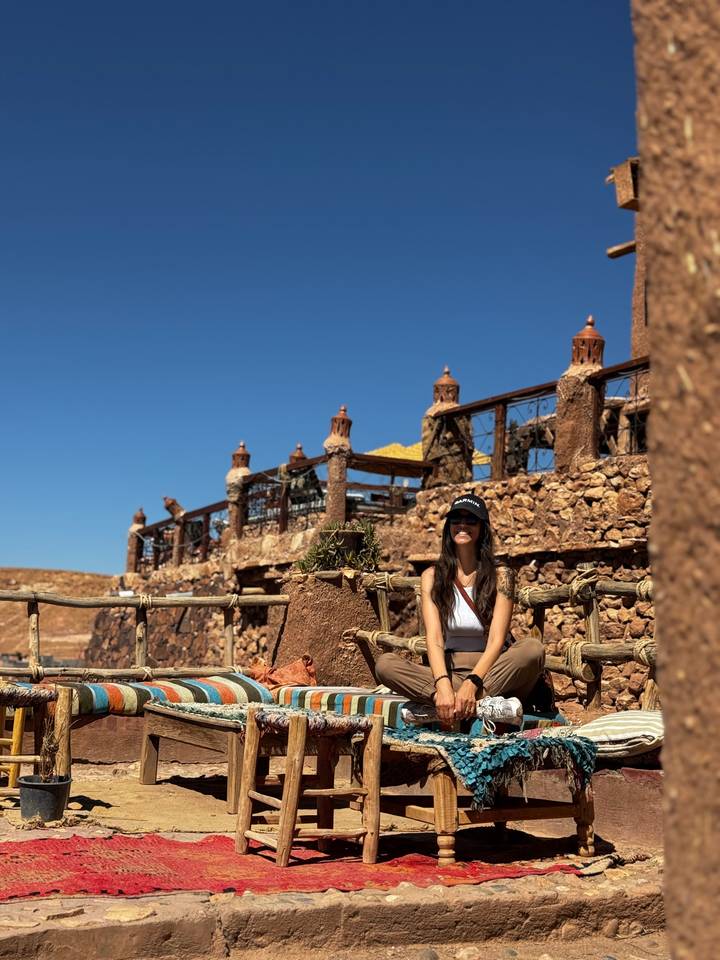 Woman in cap relaxing on rustic seating in a stone desert camp under clear blue sky.