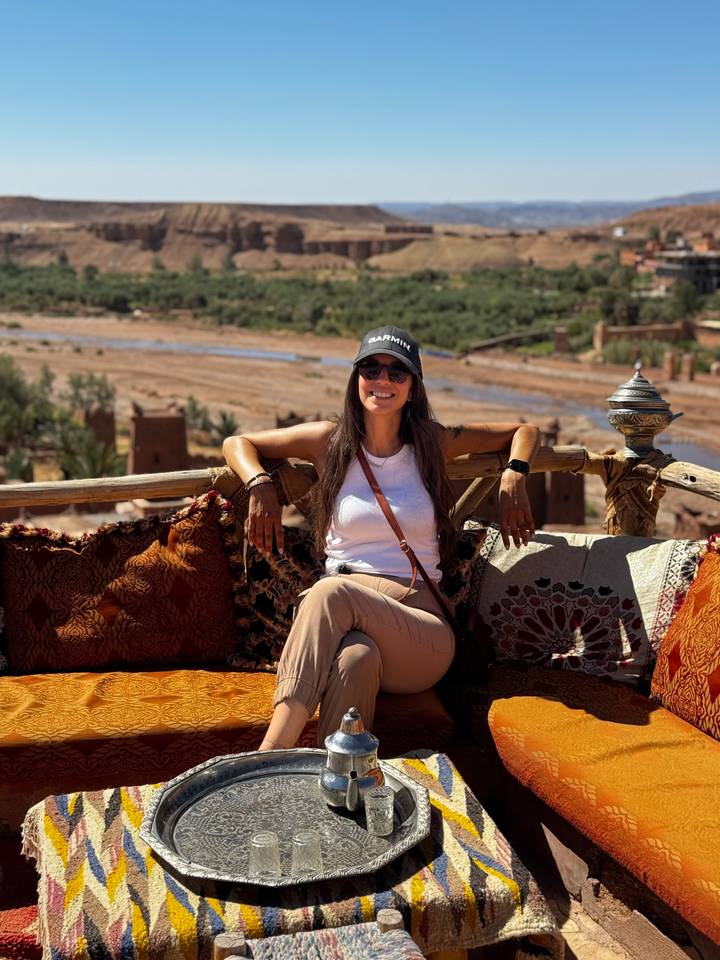 Traveller sitting on colorful cushions overlooking a river valley dotted with earthen kasbahs.