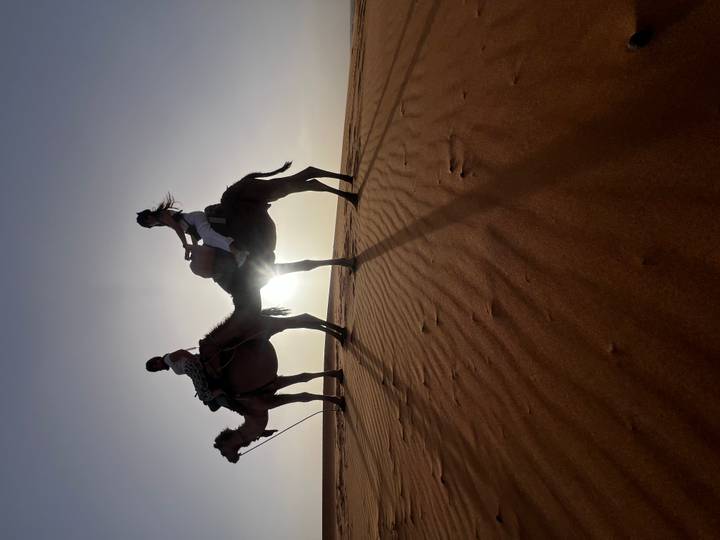 Silhouettes of two riders on camels crossing golden dunes with the sun low on the horizon.