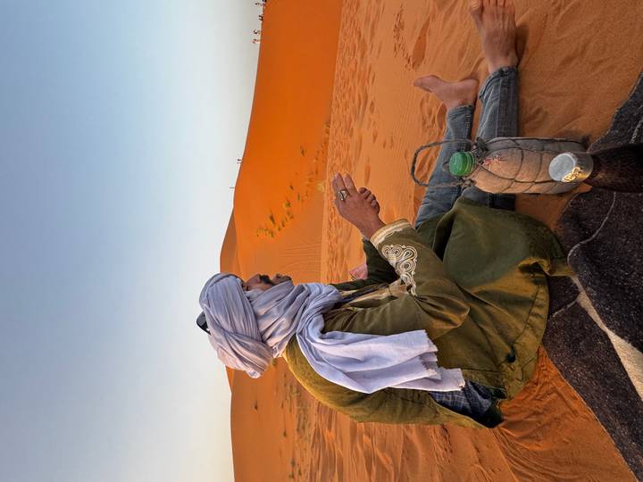 Berber man in traditional attire sitting on a dune enjoying tea at sunrise.