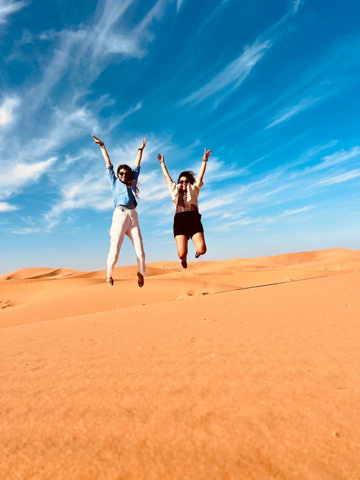 Two travellers mid-air jumping for joy on vast orange sand dunes under bright blue sky.