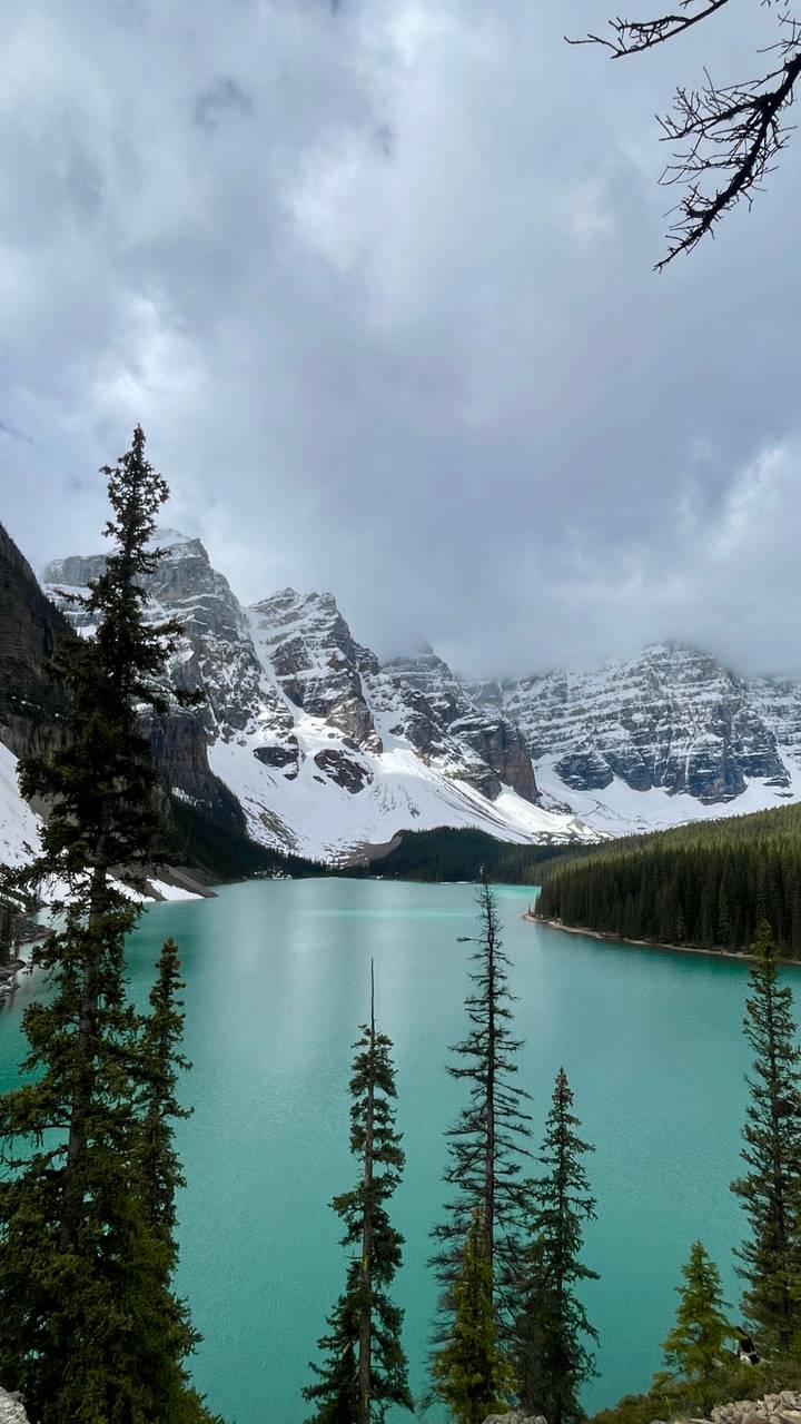 Turquoise glacial lake surrounded by snow-capped jagged peaks and evergreen forests under a dramatic cloud layer.
