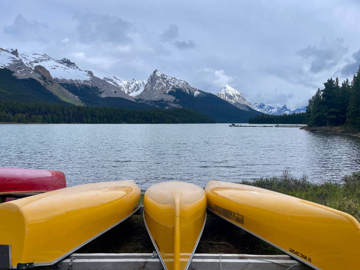 Three brightly colored canoes rest on a dock facing a broad mountain lake with dramatic snow-covered summits beyond.