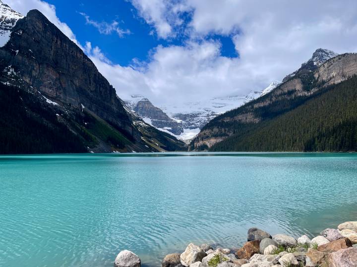 Crystal-blue Lake Louise stretches toward towering cliffs and distant glaciers beneath a patchy blue sky.