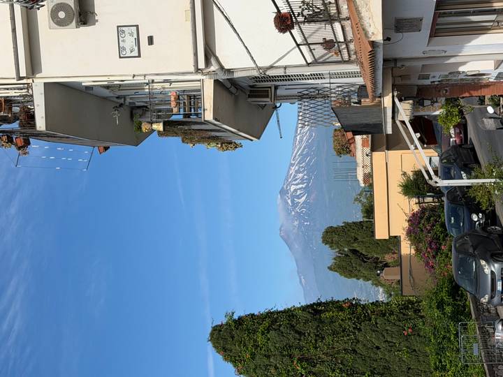 Snow-capped Mount Etna rises beyond a charming Sicilian street lined with balconies and flowers.
