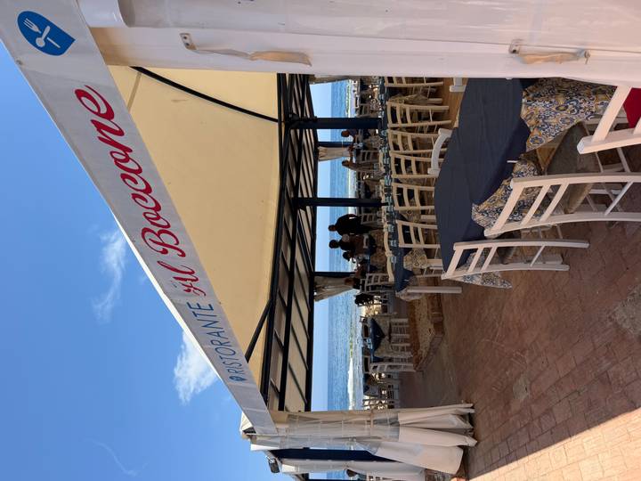 Open-air seaside restaurant with white chairs and a canvas roof overlooks gentle Mediterranean waves.